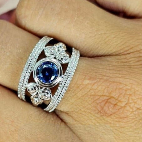A sterling silver ring with a floral filigree design and a blue cubic zirconia center, worn on a person's hand.