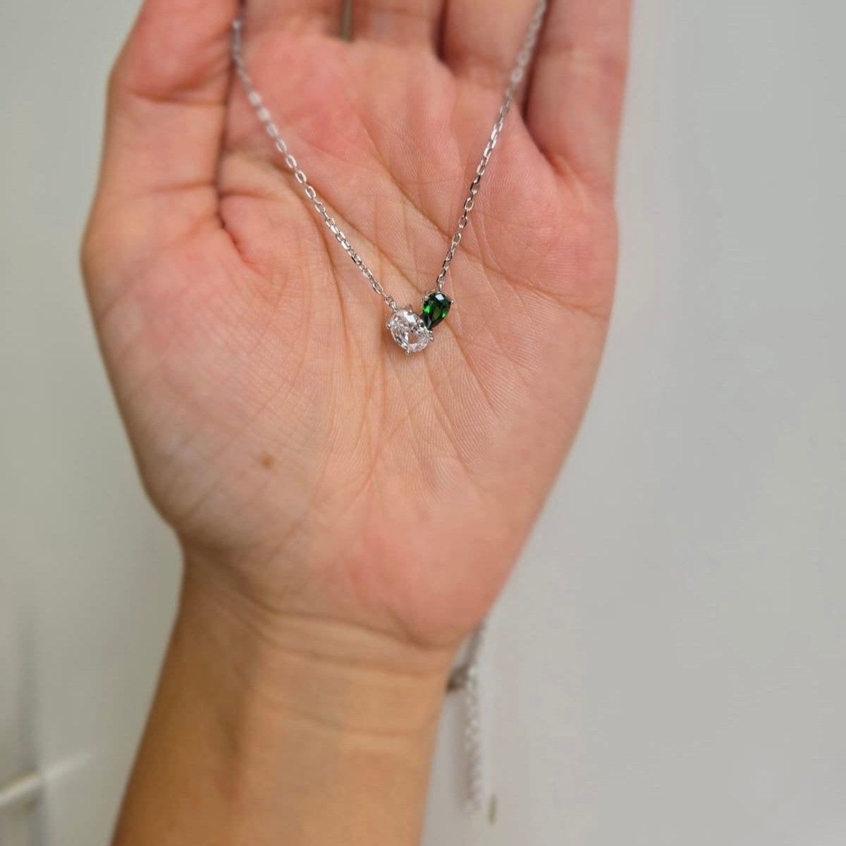 A hand holding a green and white heart-shaped necklace with cubic zirconia stones on a silver chain.