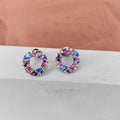 Pair of round earrings with colorful gemstones on a light gray surface.
