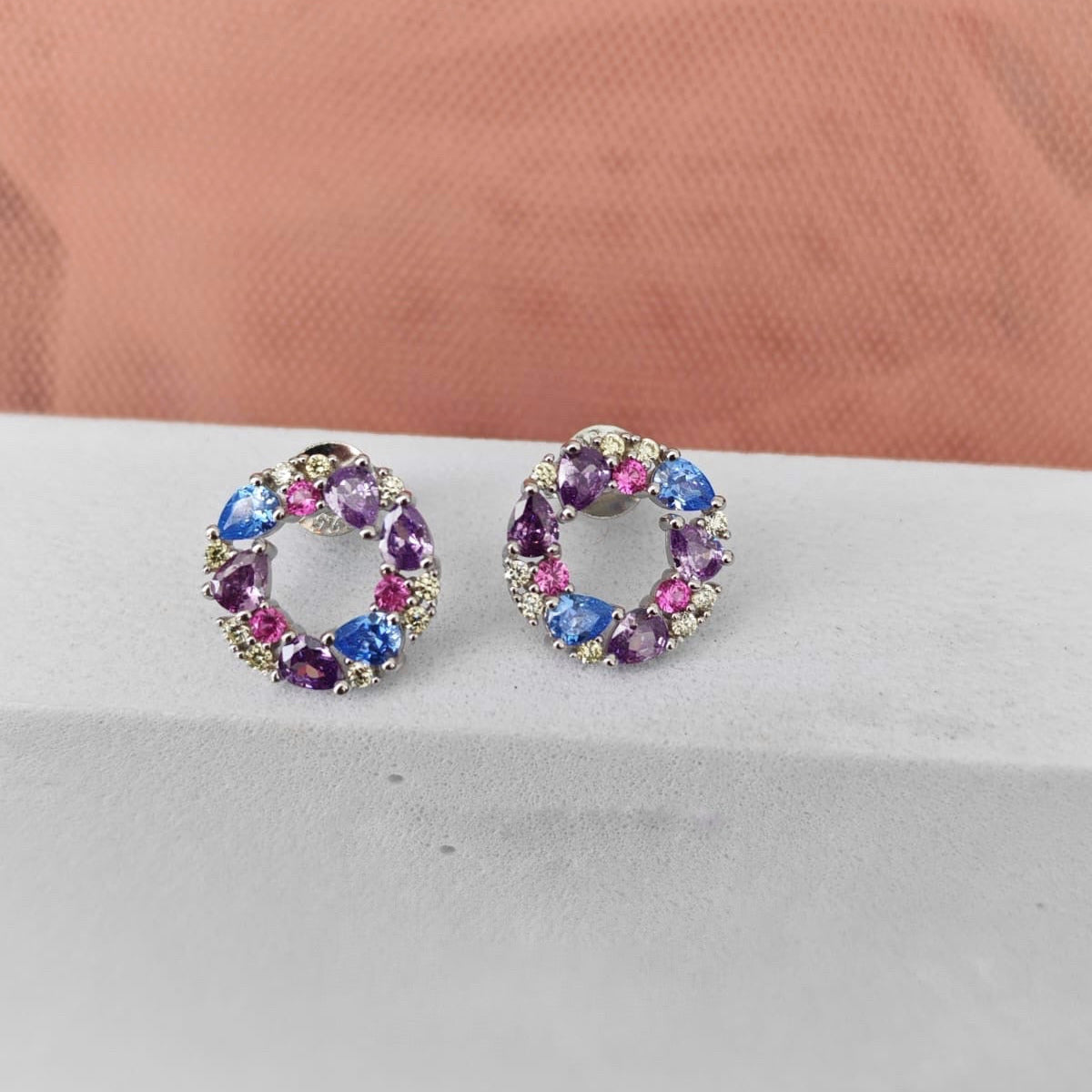 Pair of round earrings with colorful gemstones on a light gray surface.