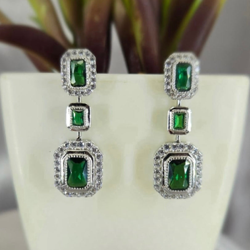 A pair of triple green drop earrings with sparkling white cubic zirconia stones, displayed against a white background with a green plant.
