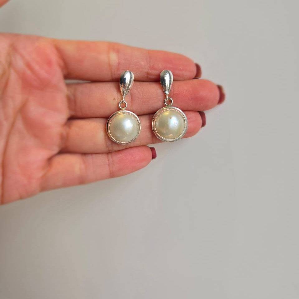 A pair of round Mabe pearl earrings with silver stem held in a person's hand.