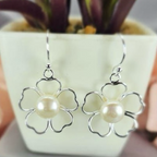A pair of sterling silver drop earrings featuring a floral design with freshwater pearl centers, displayed in front of a potted plant.