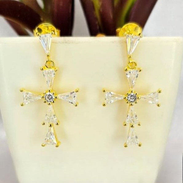 Gold cross earrings with clear stones on a white surface with green plants in the background
