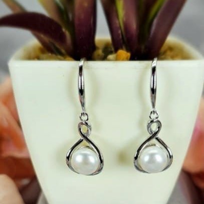 A pair of sterling silver drop earrings with freshwater pearl in an infinity hoop design, displayed on a white surface next to a potted plant.