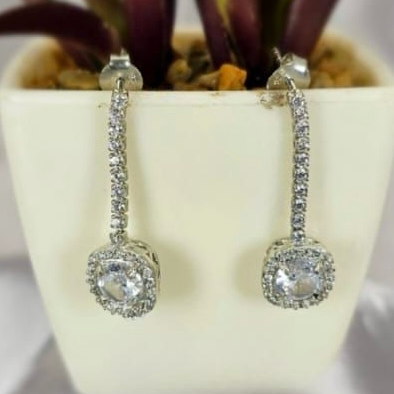 A pair of long drop earrings with cubic zirconia squares, displayed in a white vase with a green plant background.