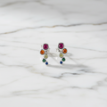 A pair of multicolored round stud earrings made with cubic zirconia, displayed in front of a white potted plant.