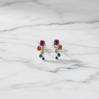 A pair of multicolored round stud earrings made with cubic zirconia, displayed in front of a white potted plant.