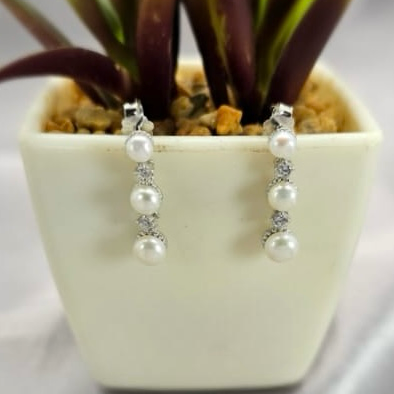 A pair of silver and pearl drop earrings displayed in a white vase with a green plant in the background.