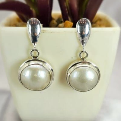 A pair of silver-colored round Mabe pearl earrings with classic silver stems, displayed in a white vase with a green plant in the background.