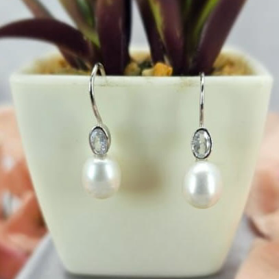 A pair of sterling silver earrings featuring freshwater pearl drops and oval cubic zirconia, displayed in a white vase with a green plant.