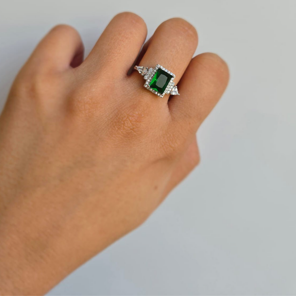 A sterling silver ring with a rectangular green cubic zirconia centerpiece and smaller white stones around it, displayed on a person's finger.