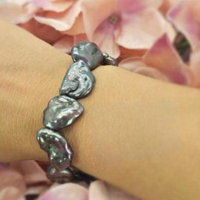 A flat, dark grey elastic bracelet with freshwater pearl accents, displayed on a wrist against a backdrop of pink flower petals.