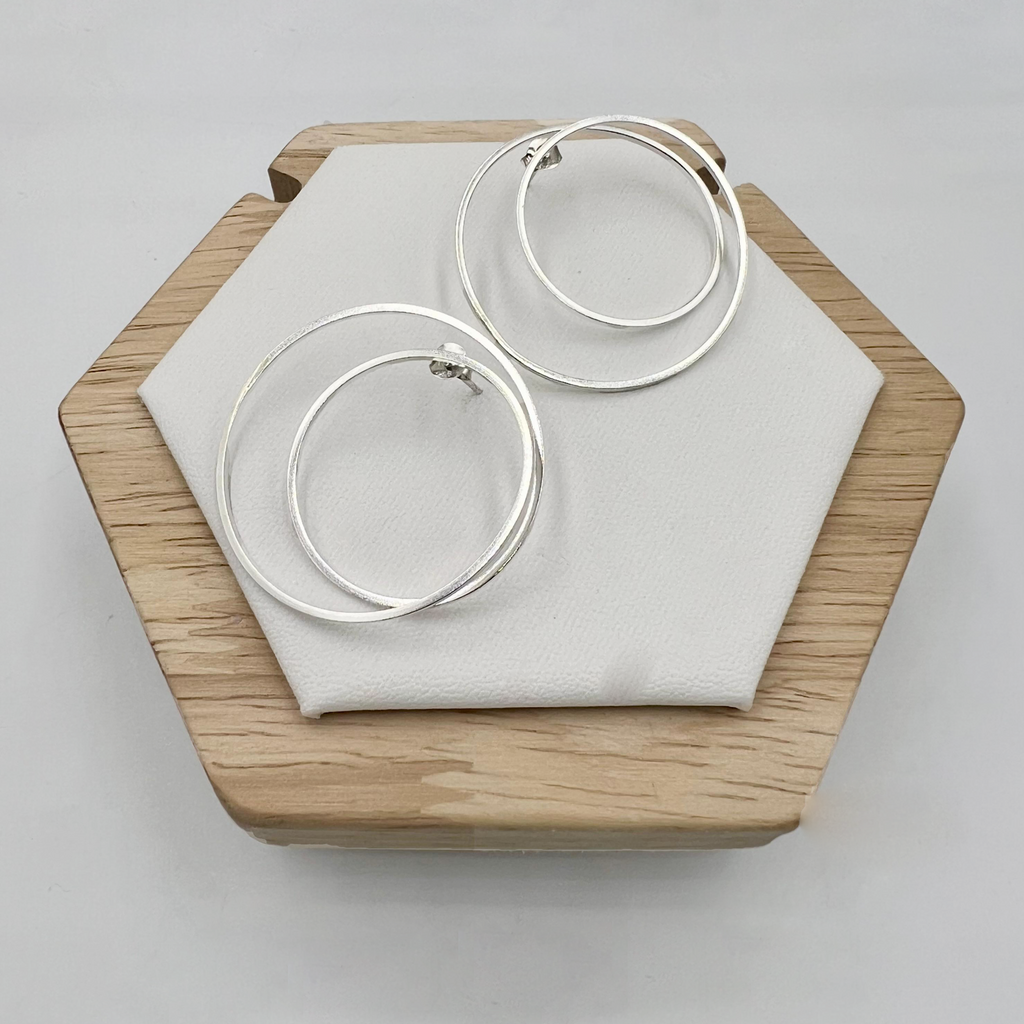A pair of large sterling silver double hoop earrings displayed on a wooden stand with a white cushion.