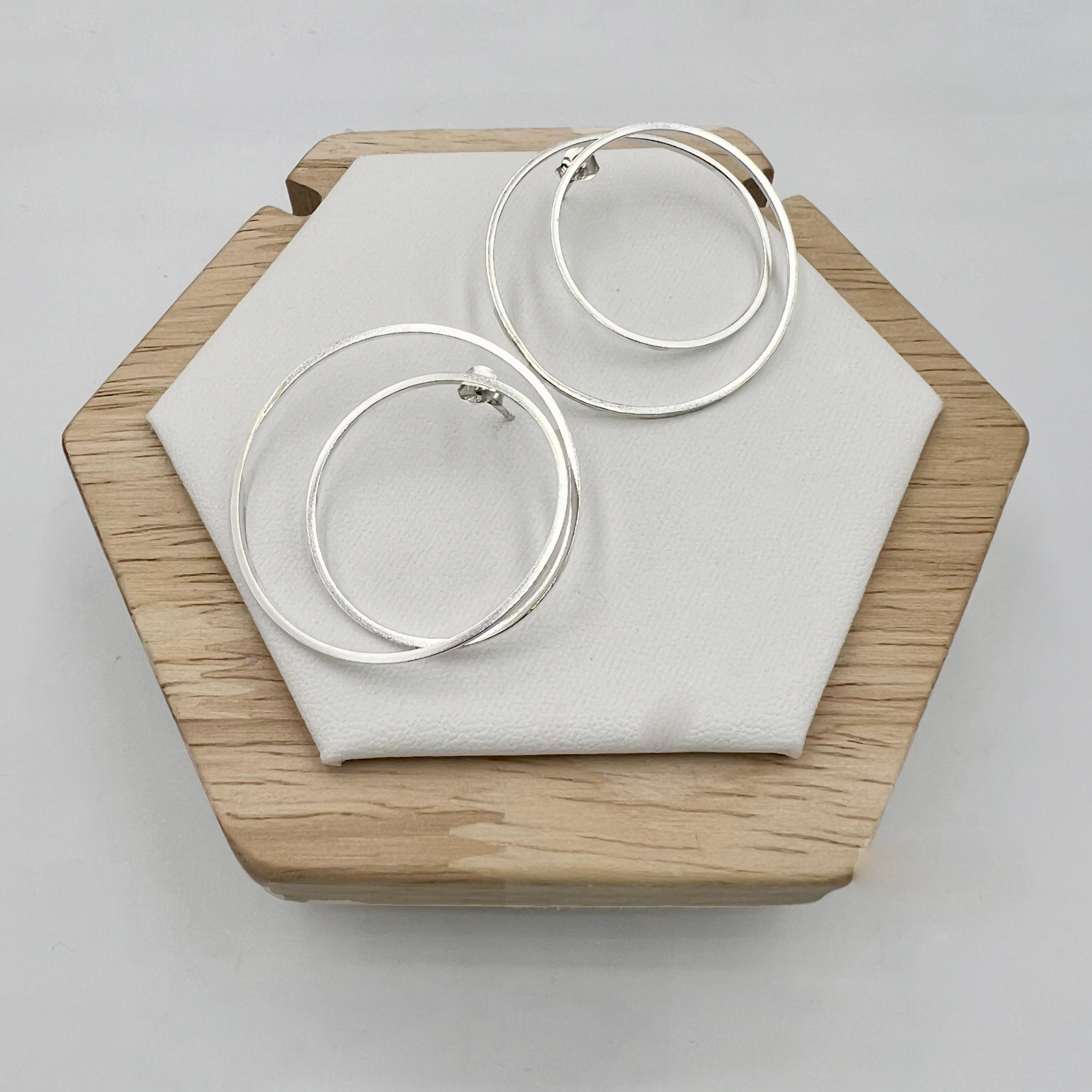 A pair of large sterling silver double hoop earrings displayed on a wooden stand with a white cushion.