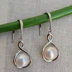 Silver earrings with pearl-like stones hanging on a green stick against a textured gray background