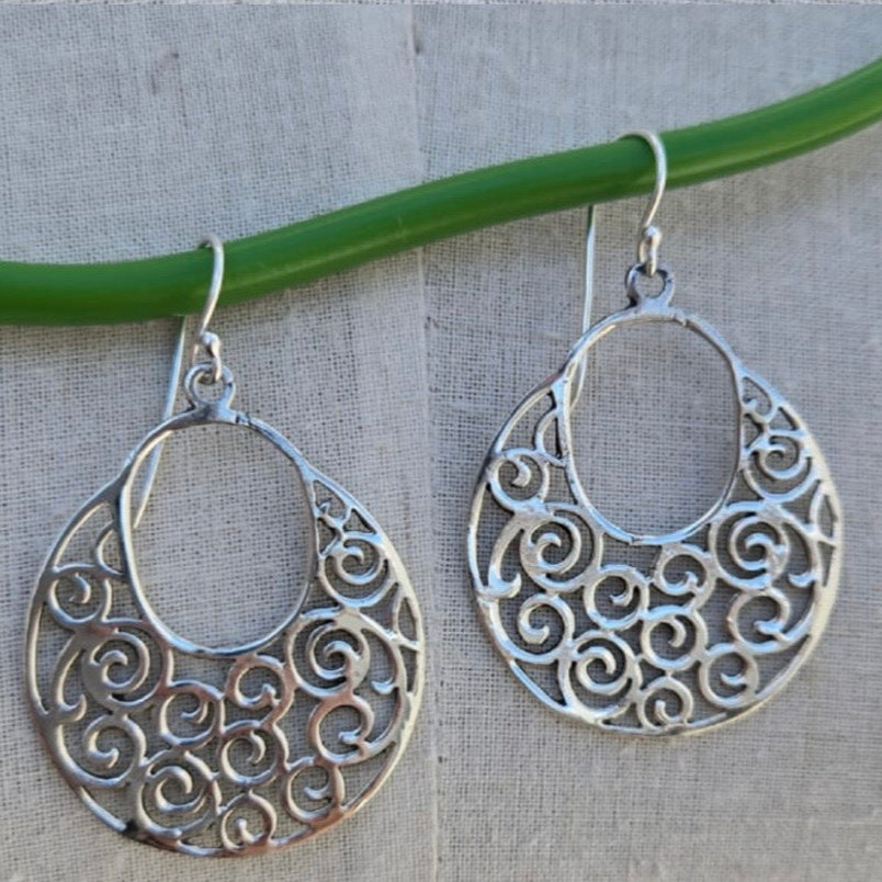 Silver hoop earrings with intricate patterns hanging on a green wire against a textured fabric background.