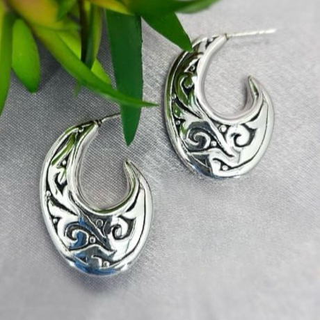 A pair of sterling silver filigree oval hoop earrings displayed on a white fabric surface with a green foliage background.