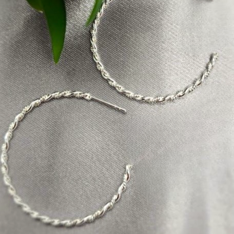 A pair of sterling silver rope hoop earrings placed on a linen surface with a green leaf and a part of a flower visible in the background.