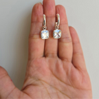 A pair of square-shaped cubic zirconia earrings attached to sterling silver hoops, displayed on a person's hand.