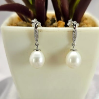 A pair of drop pearl earrings with silver hooks, displayed in a white vase with a green plant in the background.