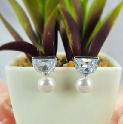 A pair of freshwater pearl drop earrings displayed on a half circle cubic zirconia top inside a white pot with green plants.