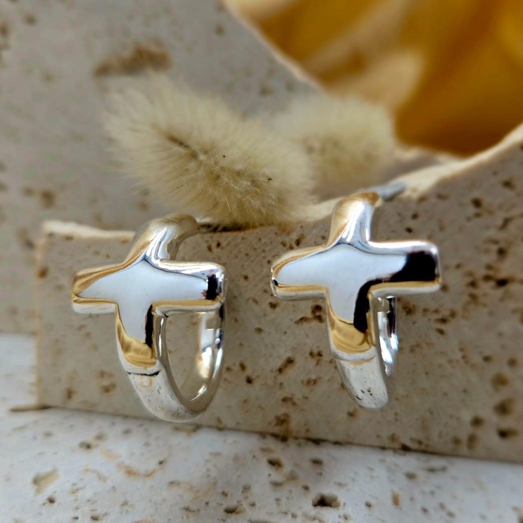 Silver cross-shaped earrings on a textured beige surface with a blurred shell in the background.