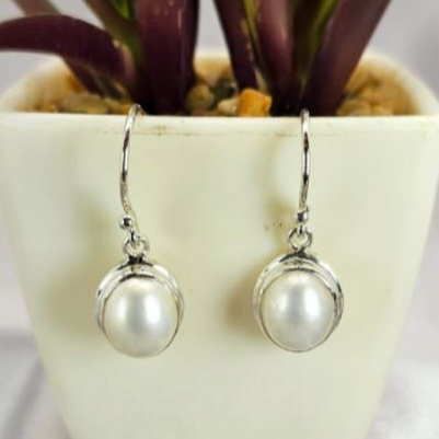 A pair of sterling silver Mabe drop earrings featuring oval pearls, displayed in front of a potted plant.