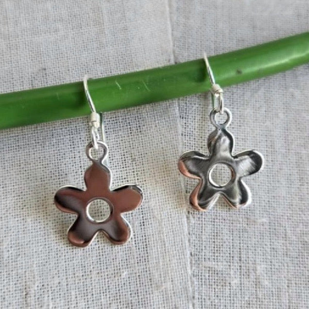 Flower-shaped silver and copper earrings on a green stick against a textured fabric background