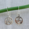 Silver triskele earrings hanging on a green rod against a light fabric background