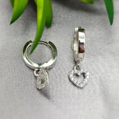Silver hoop earrings with heart and clear stone charms on a gray surface with green leaves in the background.