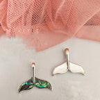 Mermaid tail earrings on a white surface with pink fabric in the background
