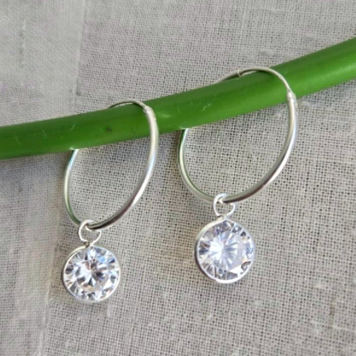 Silver hoop earrings with crystal drops on a green stick against a textured fabric background
