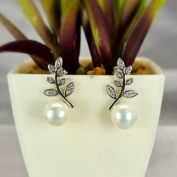 A pair of sterling silver drop earrings with cubic zirconia inset leaves and freshwater pearl drops, displayed in a white vase with green leaves.