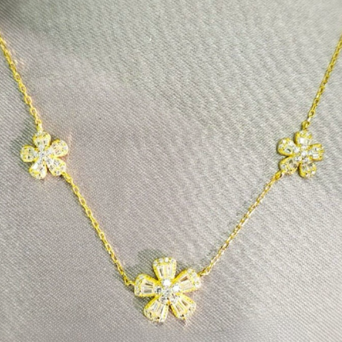 A gold necklace with three flower pendants, each adorned with what appears to be cubic zirconia stones, displayed against a grey background.