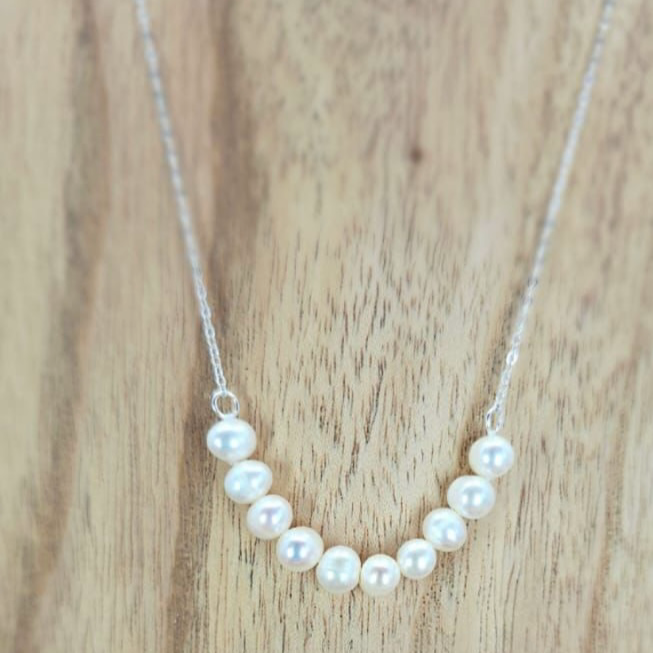A silver necklace with a semi-circular arrangement of freshwater pearls as the centerpiece, displayed against a wooden background with flowers at the bottom.