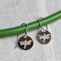 Dragonfly-themed silver earrings on a green stick against a textured fabric background
