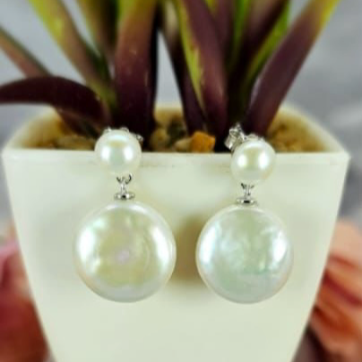 A pair of sterling silver earrings featuring a freshwater pearl drop on a coin pearl, displayed against a plant backdrop.