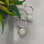 A pair of sterling silver hoop earrings with disco ball drops, displayed on a textured surface with greenery in the background.