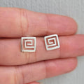 Silver square earrings held between fingers on a neutral background