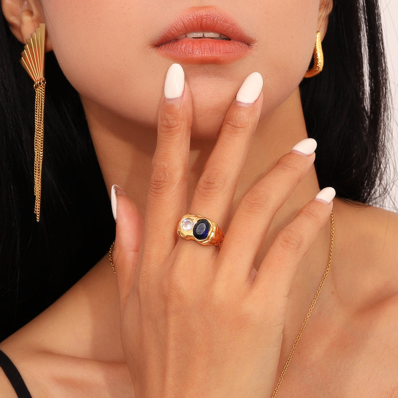 Close-up of a woman's hand wearing a gold ring with a blue gemstone, gold earrings, and white nail polish.