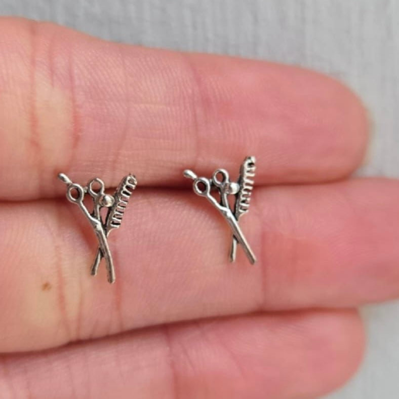 Two silver hairdresser charm pendants on a hand against a neutral background