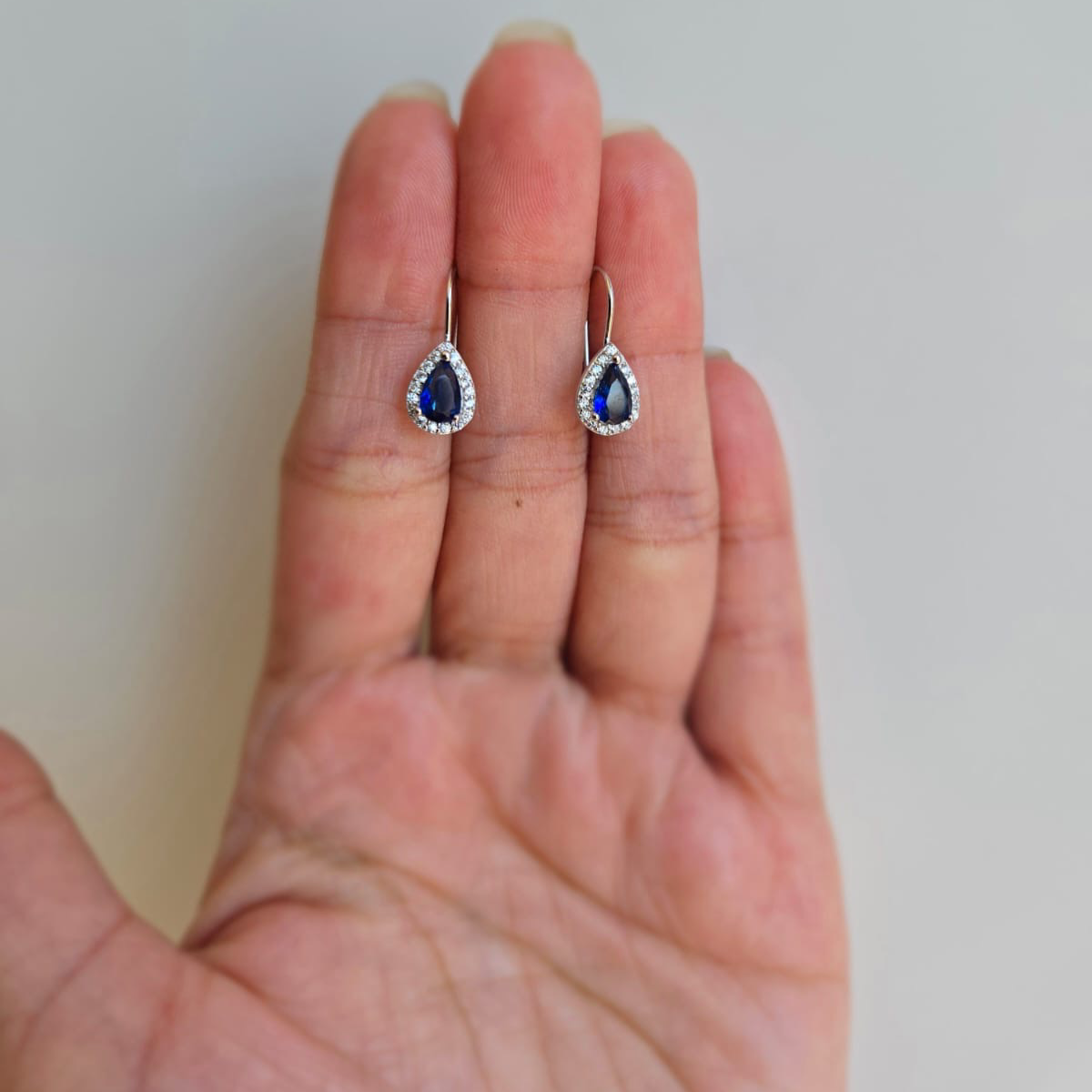 A pair of blue teardrop-shaped earrings with cubic zirconia, displayed on a person's hand.
