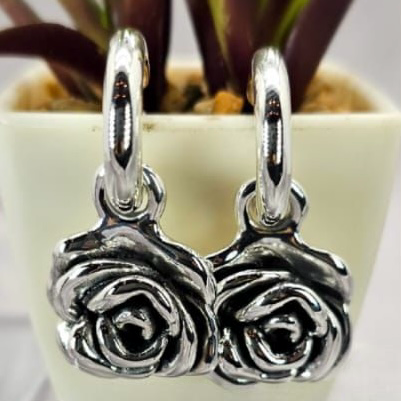 The image shows a pair of sterling silver hoop earrings with rose charms, displayed on a table next to a small potted plant.