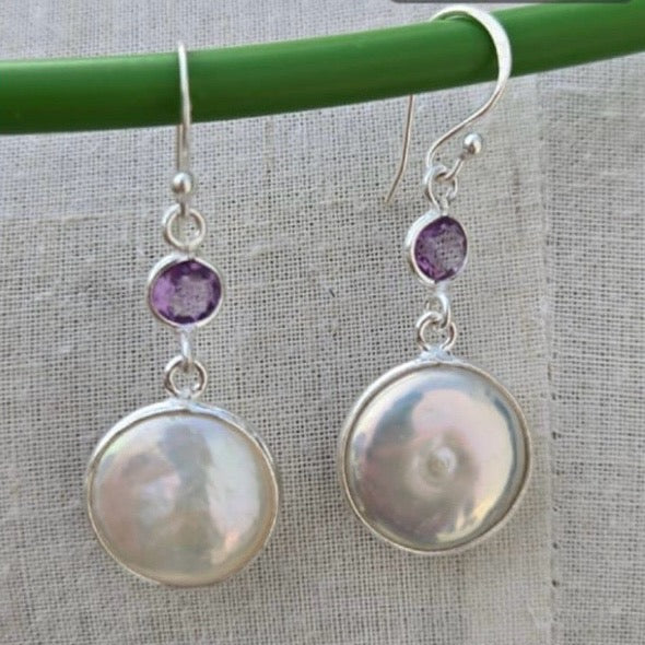 Pair of earrings with round pearl-like stones and purple gemstones on a green stick against a gray fabric background.