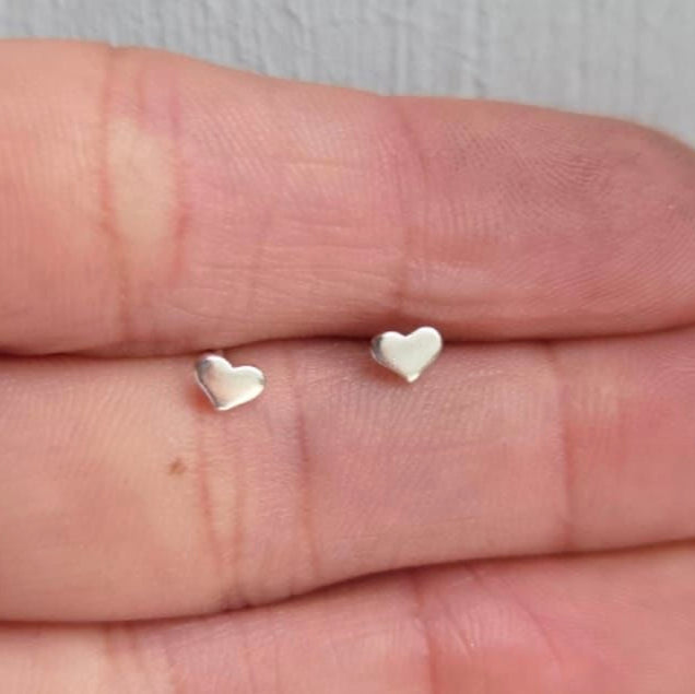 Two small silver heart-shaped earrings on a person's hand.