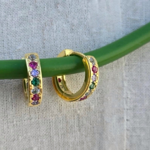 Gold hoop earrings with colorful gemstones on a green stick against a textured gray background