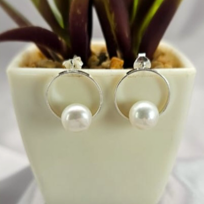 A pair of modern pearl stud earrings in silver, displayed in a white pot with green leaves.