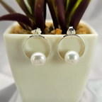 A pair of modern pearl stud earrings in silver, displayed in a white pot with green leaves.