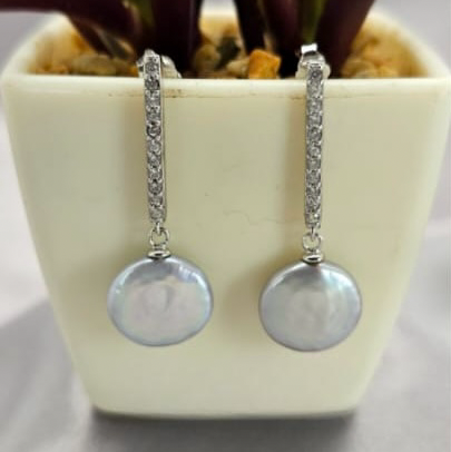 A pair of sterling silver earrings with cubic zirconia and freshwater coin pearls, displayed in a white vase with a green plant in the background.
